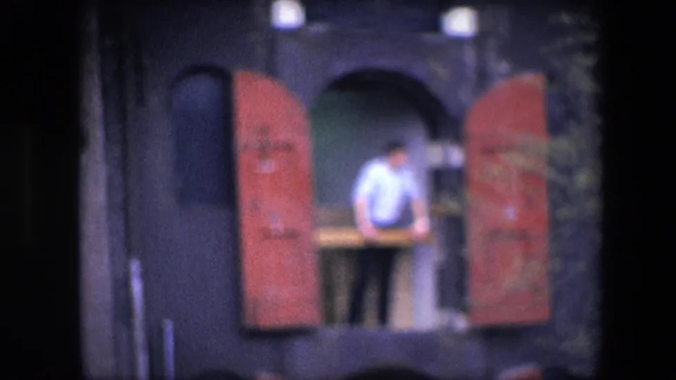 Soft 8mm frame showing the man leaning or standing near the balcony rail, centered between red shutters on a dark facade in the Amsterdam-identified clip.
