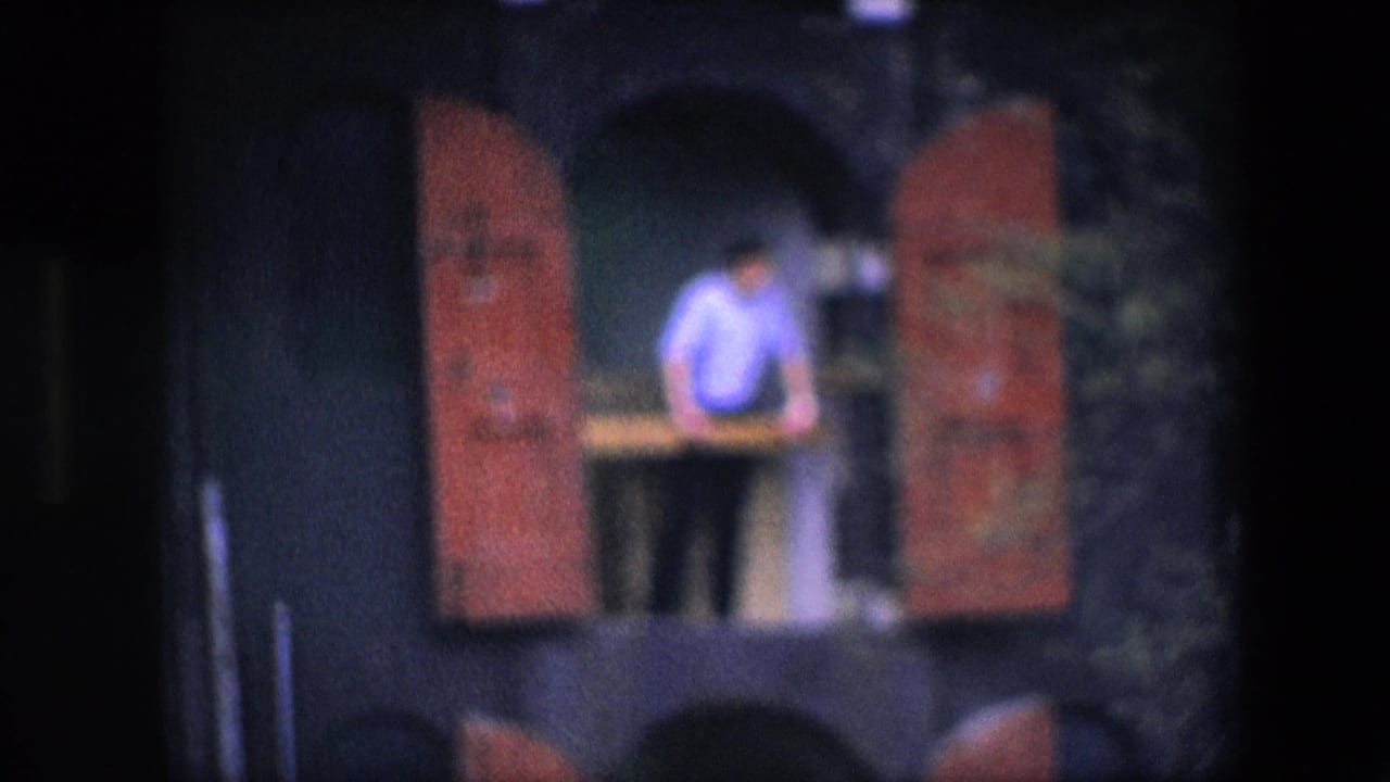 Final frame holds on the balcony figure, red shutters, arched opening, and shadowed facade, with the subject still distant and softly rendered by the 8mm scan.