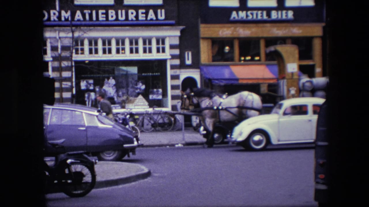 White Volkswagen Beetle passes a horse cart and parked classic car in front of storefronts.