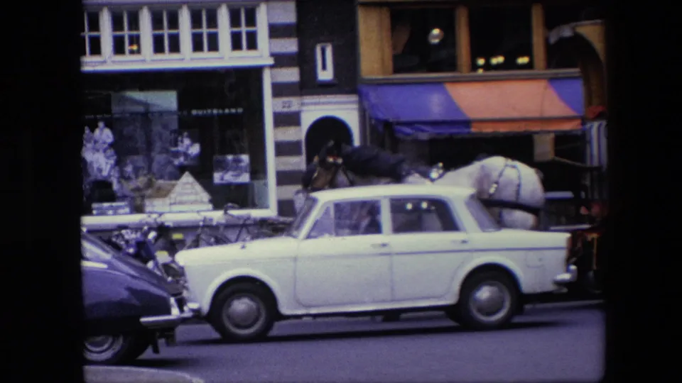 A white classic car crosses the frame in the Amsterdam street scene, partially obscuring the waiting horse cart near bicycles, pedestrians, and Dutch storefronts.