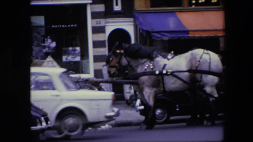 1967 Amsterdam street view with a harnessed horse and carriage passing cars in front of striped storefronts and shop windows.