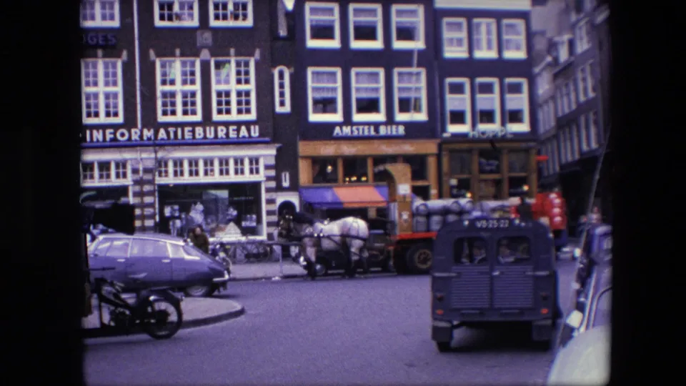 1967 Amsterdam street frame showing pedestrians near storefronts, a horse-drawn wagon loaded with barrels, a blue Volkswagen Beetle, a motorcycle, and a dark utility van in traffic.
