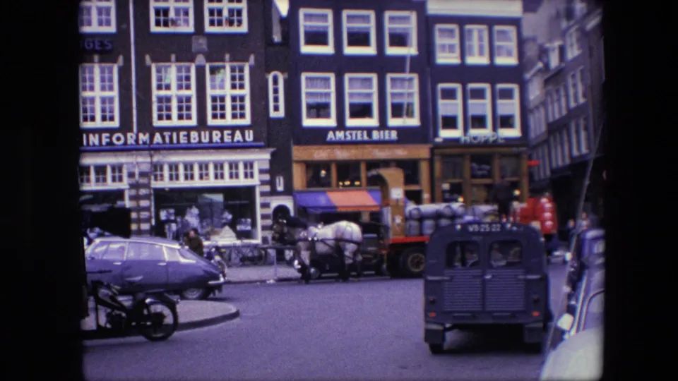 1967 Amsterdam street view with tall storefront buildings, readable INFORMATIEBUREAU and AMSTEL BIER signs, pedestrians, a horse-drawn barrel wagon, a Volkswagen Beetle, and a small utility van.