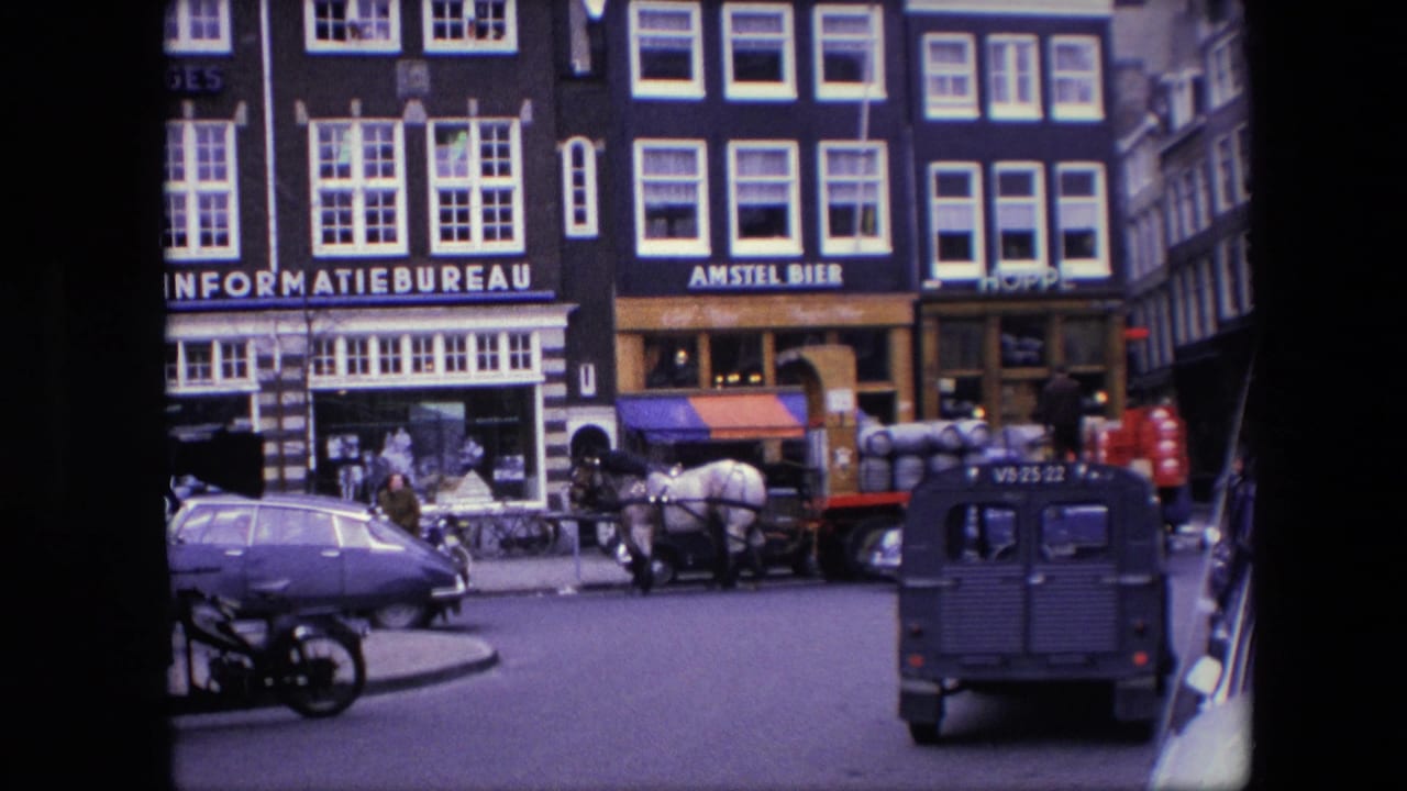 1967 Amsterdam home-movie frame showing storefront signage, a horse-drawn wagon with barrels, pedestrians on the sidewalk, a blue Volkswagen Beetle, a parked motorcycle, and a dark van on the road.