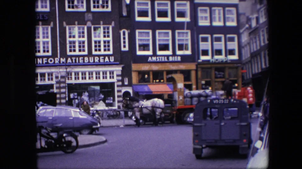 1967 Amsterdam home-movie frame showing storefront signage, a horse-drawn wagon with barrels, pedestrians on the sidewalk, a blue Volkswagen Beetle, a parked motorcycle, and a dark van on the road.