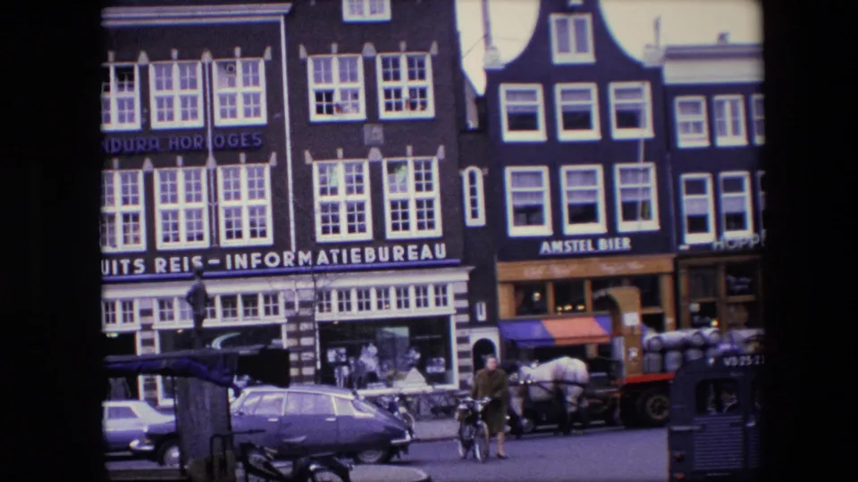 1967 Amsterdam street scene showing travel-bureau and Amstel Bier signage, a horse-drawn cart, a cyclist, pedestrians, and period vehicles.
