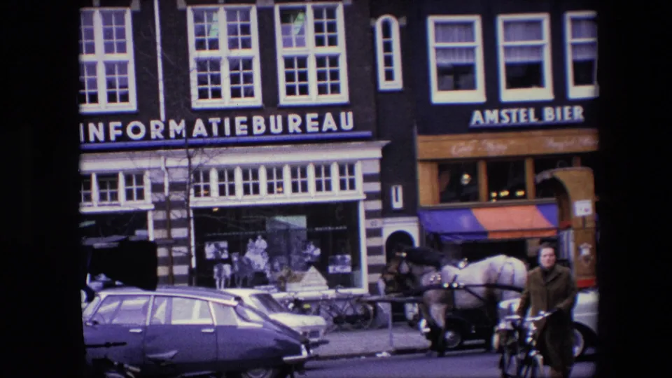 1967 Amsterdam street view showing an informatiebureau storefront, Amstel Bier signage, classic cars, bicycles, a passing cyclist, and a horse-drawn carriage.