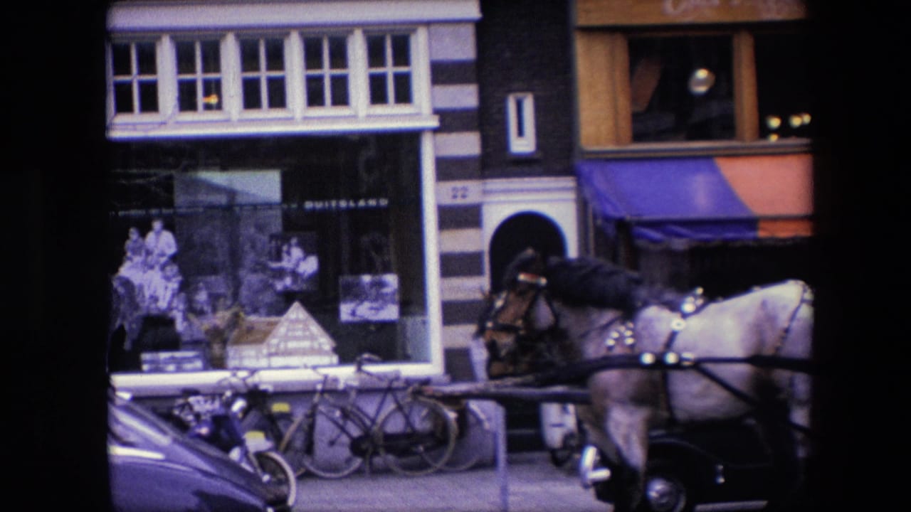 Closer 1967 Amsterdam view of curbside bicycles, a parked classic car, shop windows, striped facade details, and a carriage horse passing the Amstel Bier frontage.