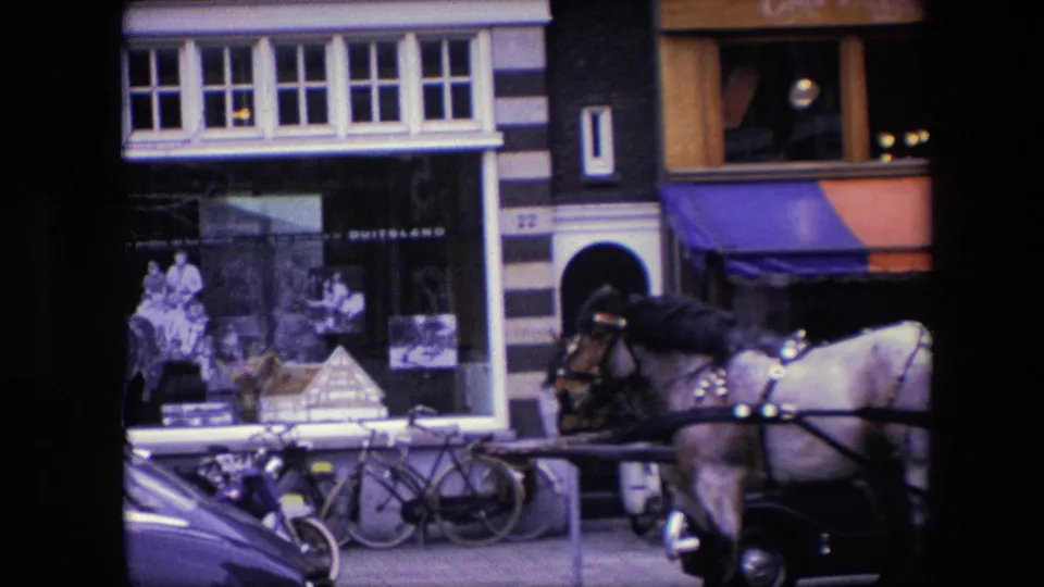 Closer 1967 Amsterdam view of curbside bicycles, a parked classic car, shop windows, striped facade details, and a carriage horse passing the Amstel Bier frontage.