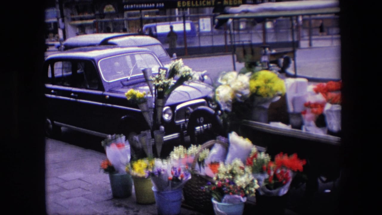 1967 Amsterdam street view with colorful flower buckets in the foreground, a dark parked car at left, and shopfront signage across the road.