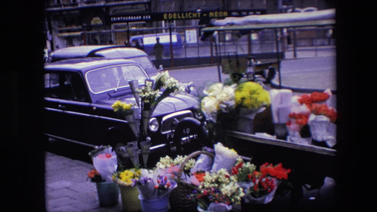 Amsterdam flower display filmed from a fixed angle, with bouquets in the foreground, a dark parked car, and visible shop signage across the street.