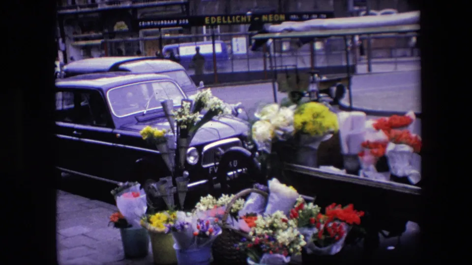Amsterdam flower display filmed from a fixed angle, with bouquets in the foreground, a dark parked car, and visible shop signage across the street.