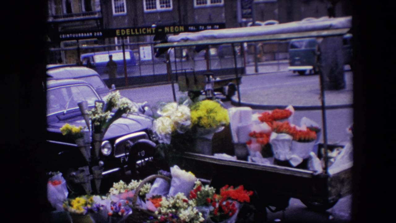 1967 Amsterdam street scene with a flower cart of red, yellow, and white blooms beside older cars and storefronts, framed by dark 8mm film borders.