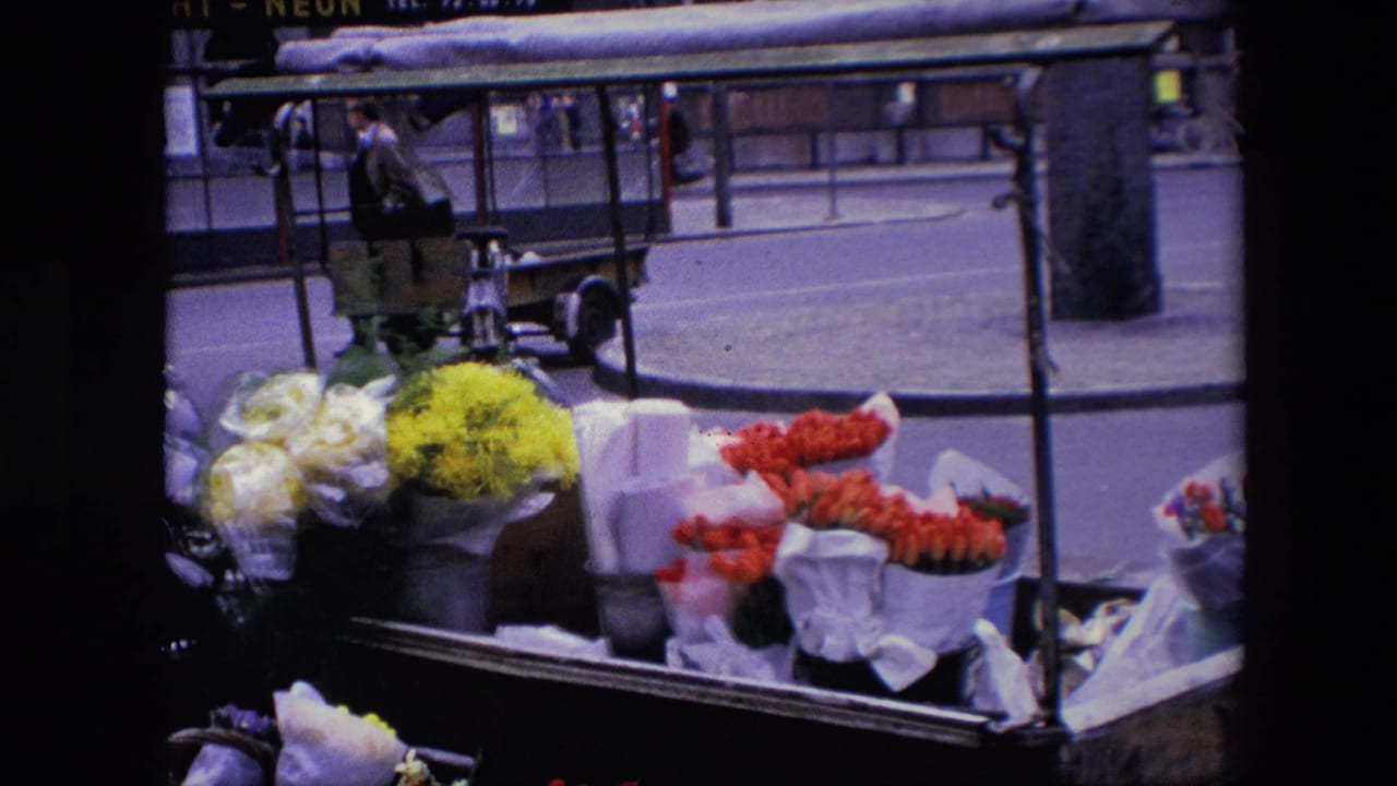 Closer 8mm view of the flower cart, with bright yellow, red, and pale wrapped bouquets filling the lower frame against a city street background.