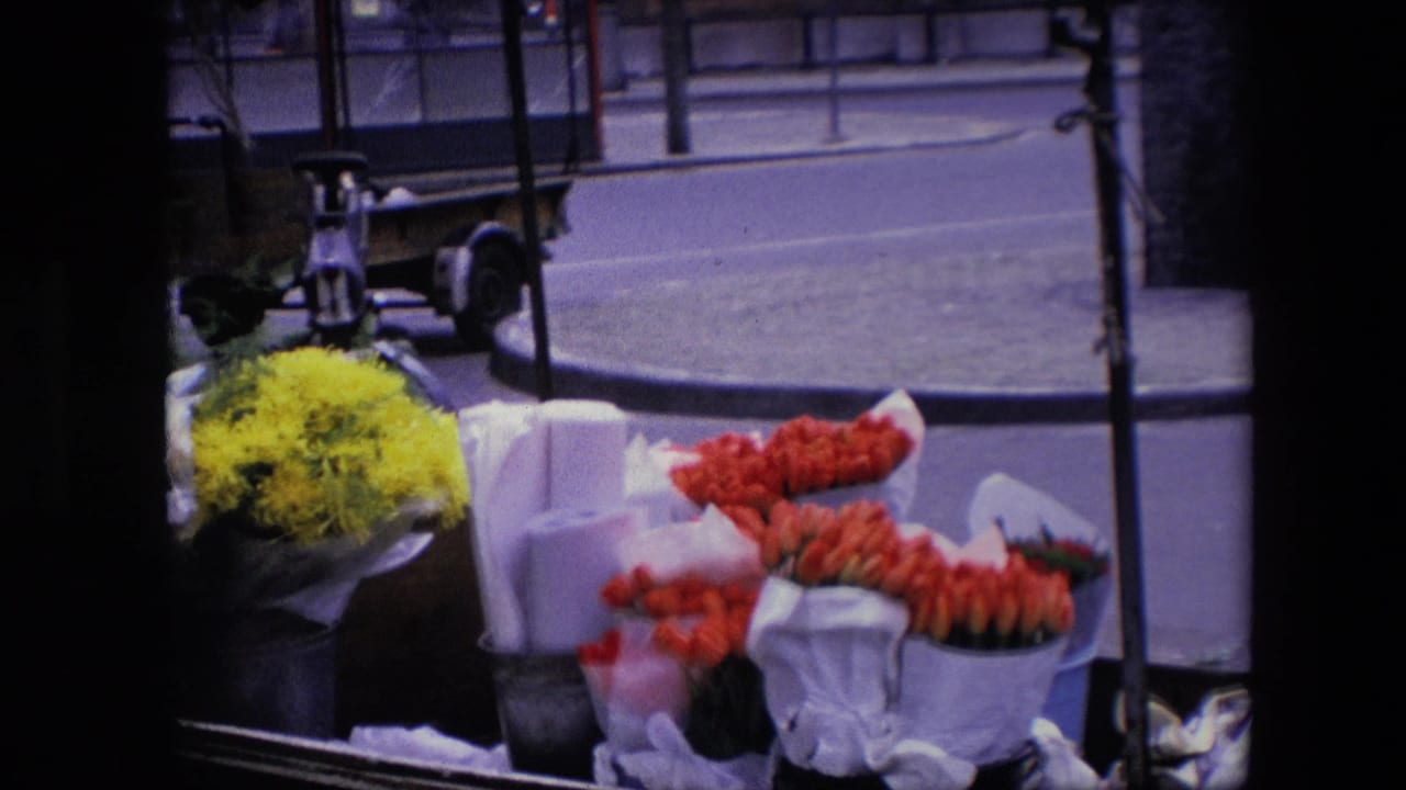 Final frame holds on the flower cart detail, showing red and yellow flowers near the curb with part of a parked vehicle and street surface behind it.
