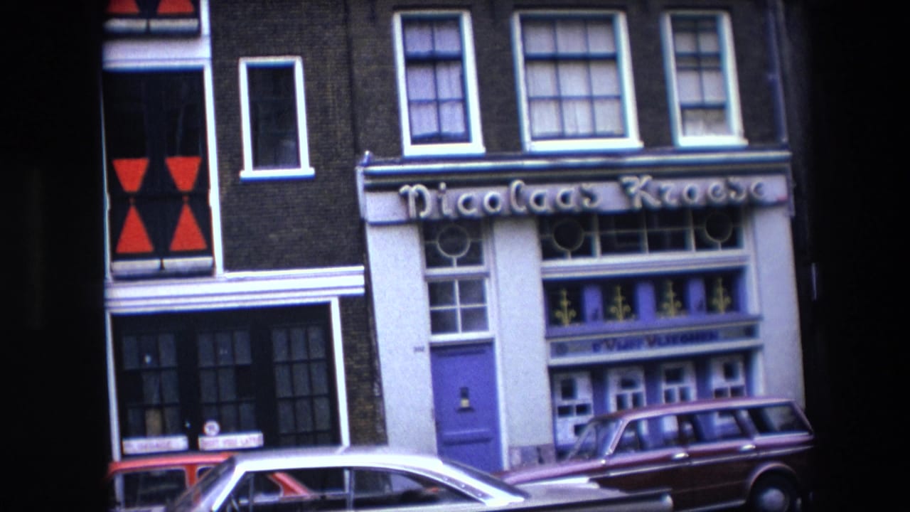 Amsterdam street facade with Nicolaas Kroese lettering, a purple door, upper windows, and parked cars, seen in 1967 8mm home-movie footage per metadata.