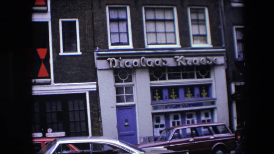 Street-level 8mm view of a building exterior with Nicolaas Kroese lettering, purple door, multiple windows, and parked cars in Amsterdam, supported by 1967 clip metadata.