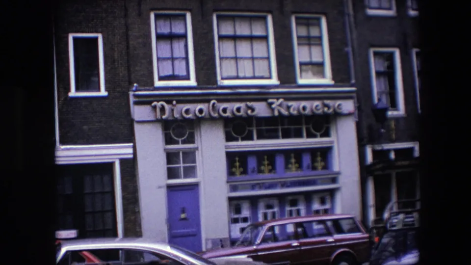 1967 Amsterdam 8mm frame showing the Nicolaas Kroese facade with a purple entrance, white window trim, dark brick upper floors, and cars parked at the curb.