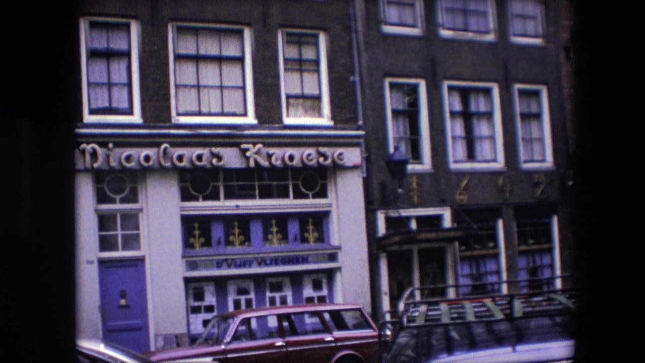 1967 Amsterdam 8mm still showing a closer portion of the Nicolaas Kroese storefront, including the sign, painted entrance, decorative glass panels, brick upper story, and parked vehicle.