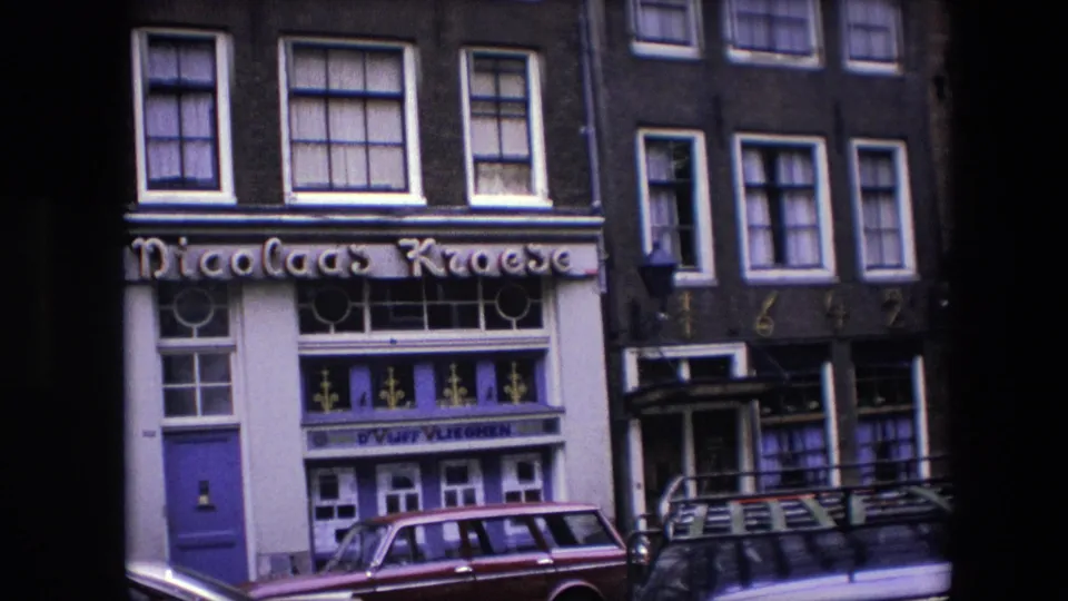 1967 Amsterdam 8mm still showing a closer portion of the Nicolaas Kroese storefront, including the sign, painted entrance, decorative glass panels, brick upper story, and parked vehicle.