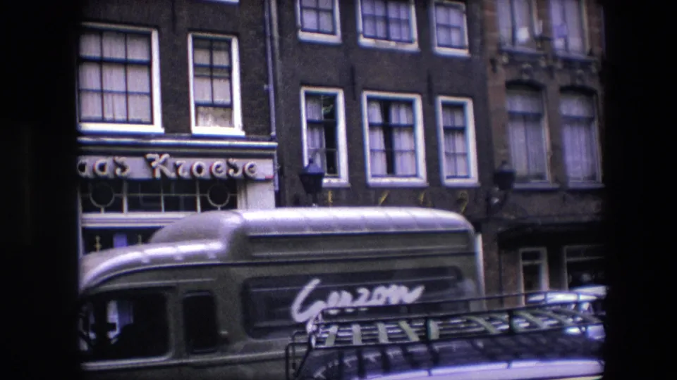 A green bus with visible lettering passes along a 1967 Amsterdam street in front of the Kroese storefront and multi-story buildings.