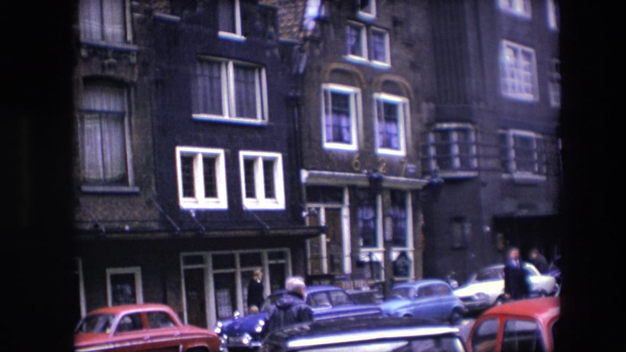 1967 Amsterdam street scene with dark narrow facades, parked compact cars, pedestrians, and tall windows below apartment-like upper floors.