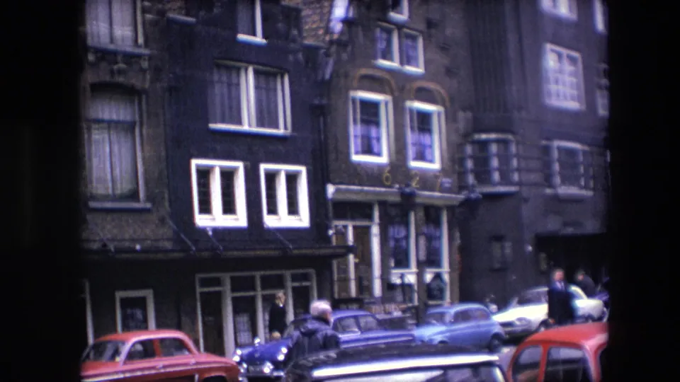 1967 Amsterdam street scene with dark narrow facades, parked compact cars, pedestrians, and tall windows below apartment-like upper floors.