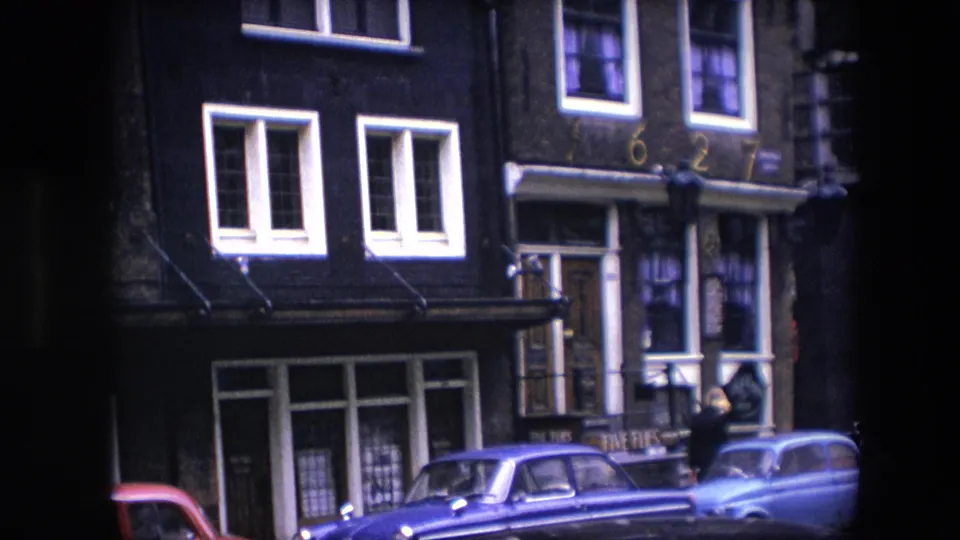 Amsterdam street view identified as 1967, showing a dark brick building with white windows, parked cars, shopfronts, and a person near the entrance.
