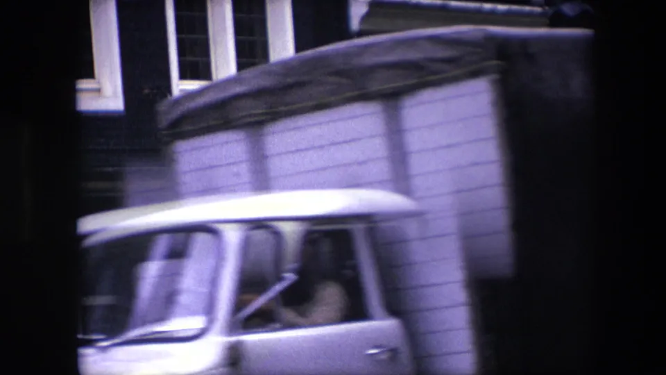 A light-colored van or truck passes across the 1967 Amsterdam street view, partially obscuring the brick building and parked cars behind it.