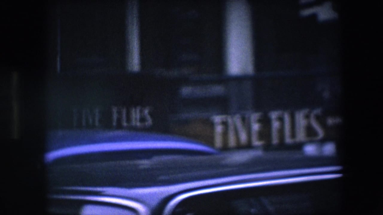 Soft, underexposed 8mm frame of the Amsterdam storefront sign and a blue car roof, with the street scene mostly obscured by dark foreground framing.
