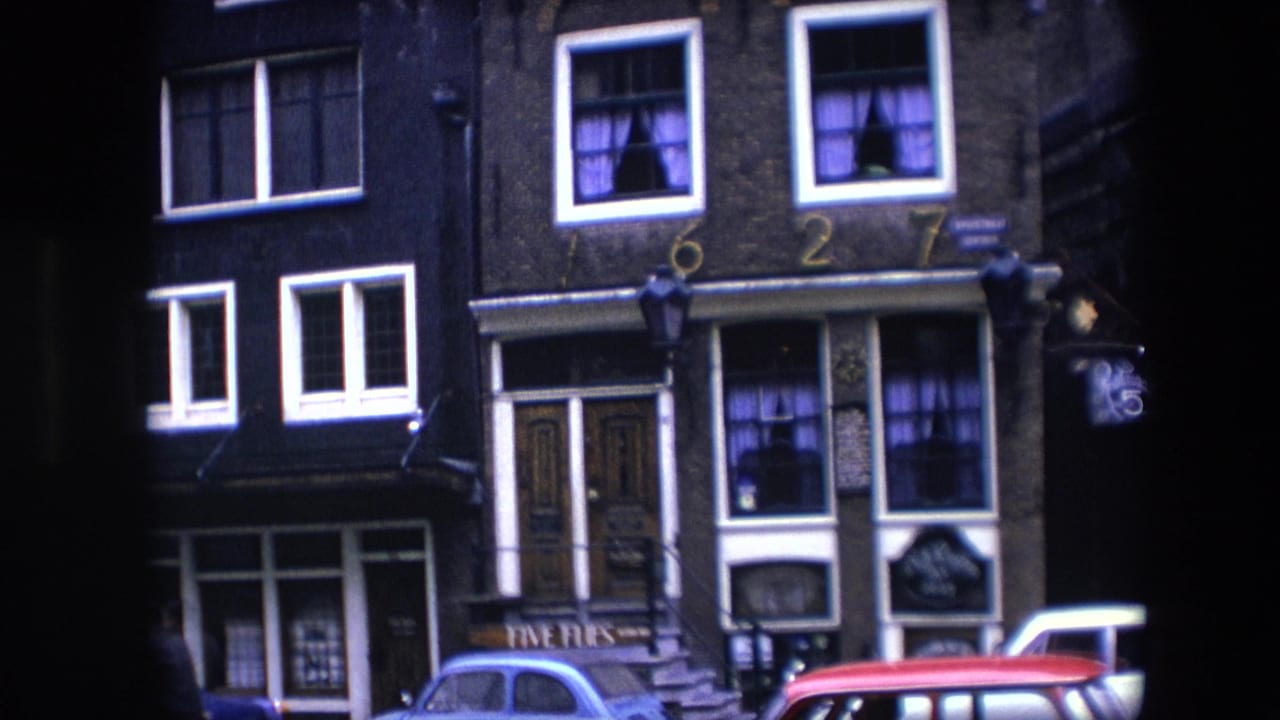 1967 Amsterdam street facade showing a brick building with white-trimmed windows, purple curtains, storefront signage, and blue, red, and white cars at the curb.