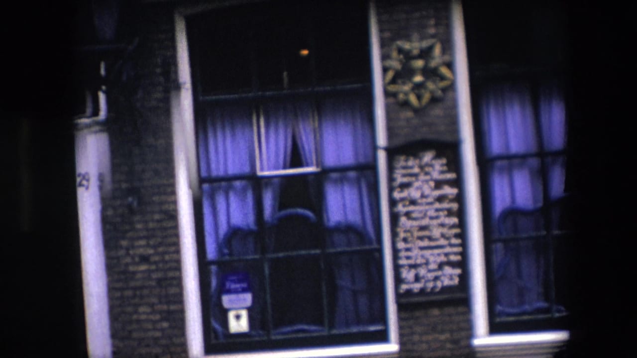 1967 Amsterdam home-movie frame showing a nighttime brick facade with purple curtains, white wall lettering, vertical pipes, and a small blue sign in a lower window.