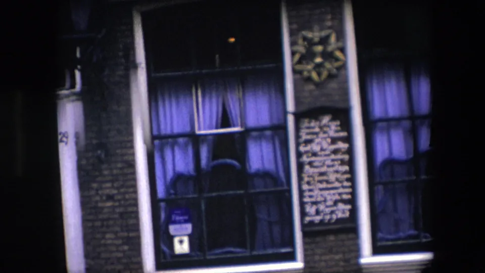 1967 Amsterdam home-movie frame showing a nighttime brick facade with purple curtains, white wall lettering, vertical pipes, and a small blue sign in a lower window.