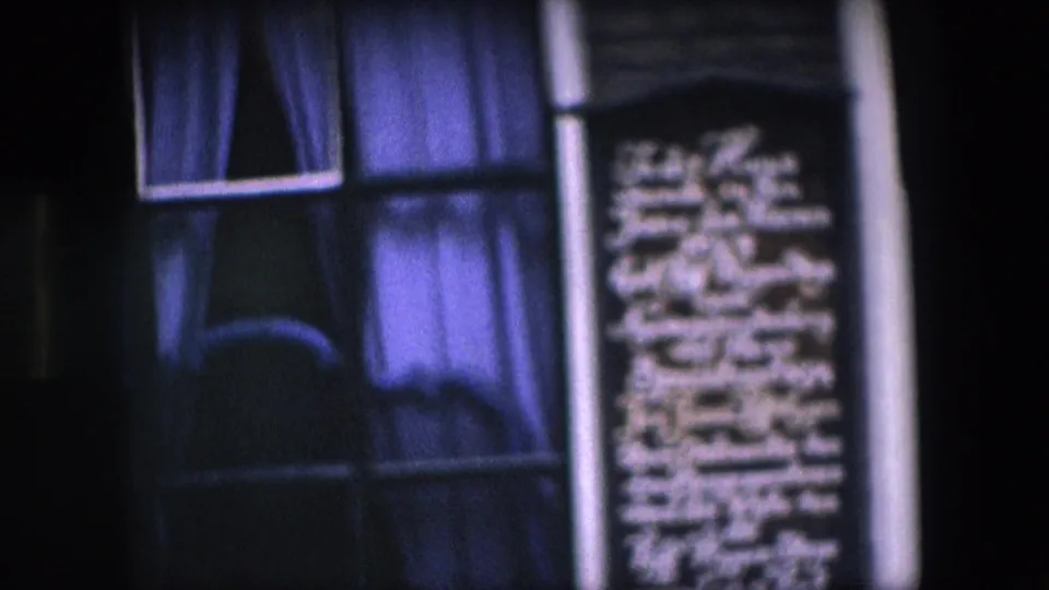 Tight, soft 8mm view of a commemorative plaque set into a dark brick Amsterdam facade between curtained windows.