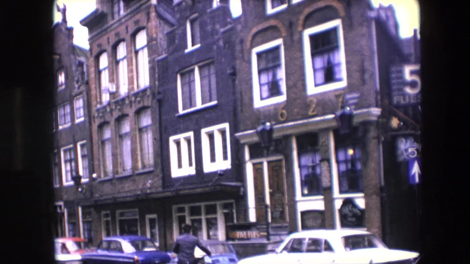 Wider 1967 Amsterdam street view with narrow brick buildings, white-trimmed windows, parked cars, shopfront signage, and a distant pedestrian in the roadway.
