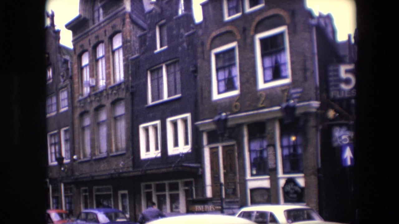 1967 Amsterdam home-movie frame showing tall brick canal-house facades, white-framed windows, parked cars, and a visible 5 FLIES corner sign through dark 8mm borders.