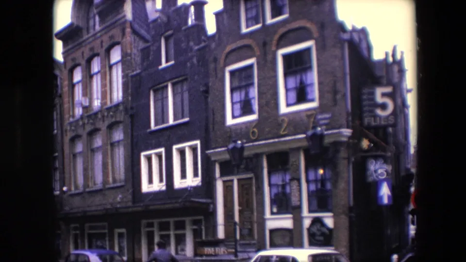 1967 Amsterdam frame with gabled canal-house facades, white-trimmed windows, parked cars along the street, and a 5 FLIES sign visible on the right.