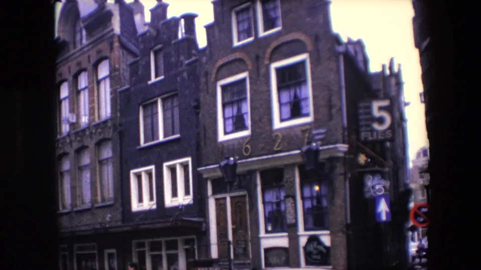 1967 Amsterdam home-movie frame showing gabled brick buildings, white-framed windows, parked cars, a small passerby near the lower edge, and the 5 FLIES sign on the corner.