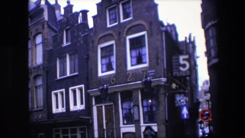 1967 Amsterdam frame showing a row of tall gabled brick houses with white-trimmed windows, parked cars below, dark 8mm borders, and a 5 FLIES sign at right.