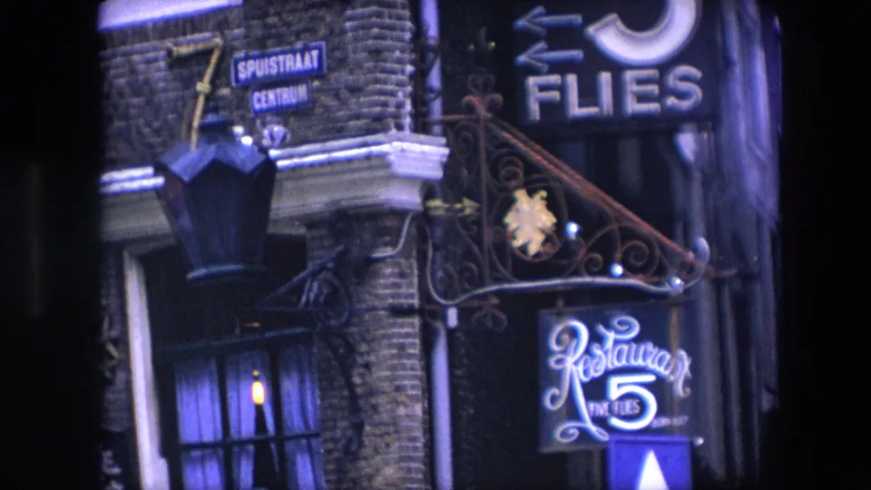 1967 8mm frame of an Amsterdam brick building entrance showing Dutch street signs, wrought iron scrollwork, and the illuminated Restaurant Five Flies sign at night.