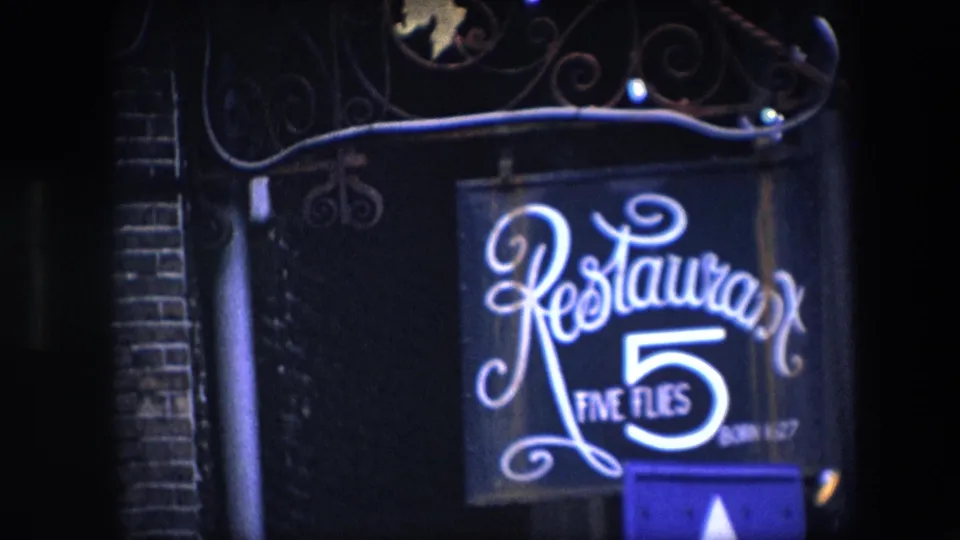 1967 8mm zoom-in frame centered on the illuminated Restaurant Five Flies sign, with ornate ironwork and brick edges visible in the night exposure.