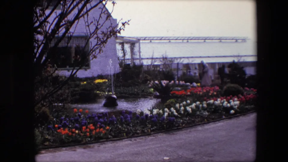 1967 8mm frame identified as Amsterdam showing a landscaped garden with a small pond, fountain jets, colorful flower beds, a path, trees, and a pale building beyond.