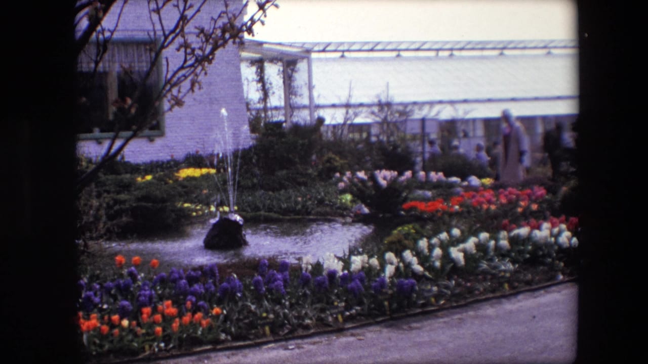 Landscaped flower garden with pond, fountain, visitors, trees, and greenhouse-like building.