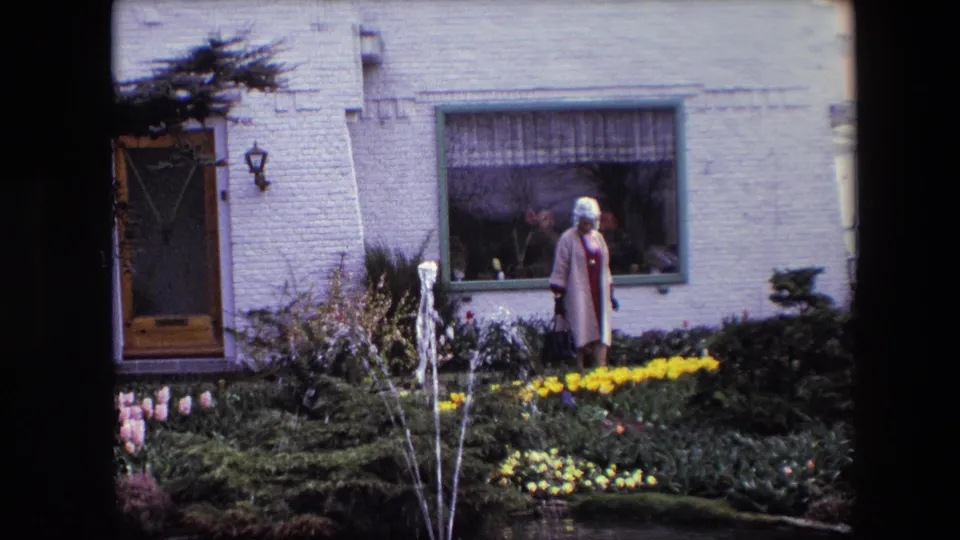 An older woman in a light coat and hat stands beside fountain jets and yellow flowers in front of a pale brick residential facade with a curtained window.