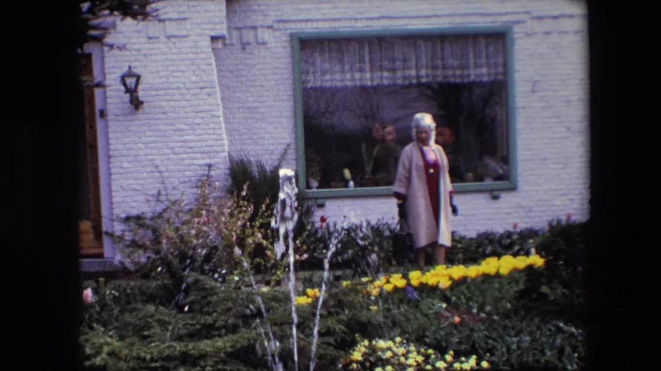 An older woman stands among flower beds near a small fountain, with a pale brick wall, curtained window, doorway edge, and garden shrubs visible behind her.