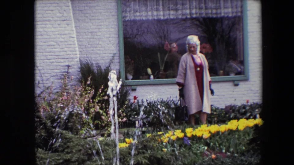 A woman in 1960s-style dress stands in a garden near yellow tulips, a small fountain, and a white brick building with a large window behind her.
