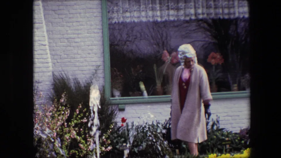 A woman in a light coat and white hat looks down as she moves through a planted garden with fountain water, spring flowers, and a large window in the background.