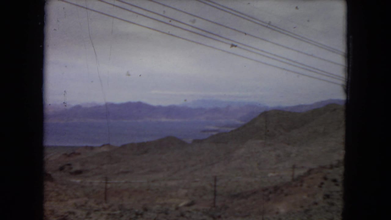 1967 United States 8mm frame showing scrubby desert hills, a distant lake or reservoir, layered mountains, fence posts, and overhead power lines.