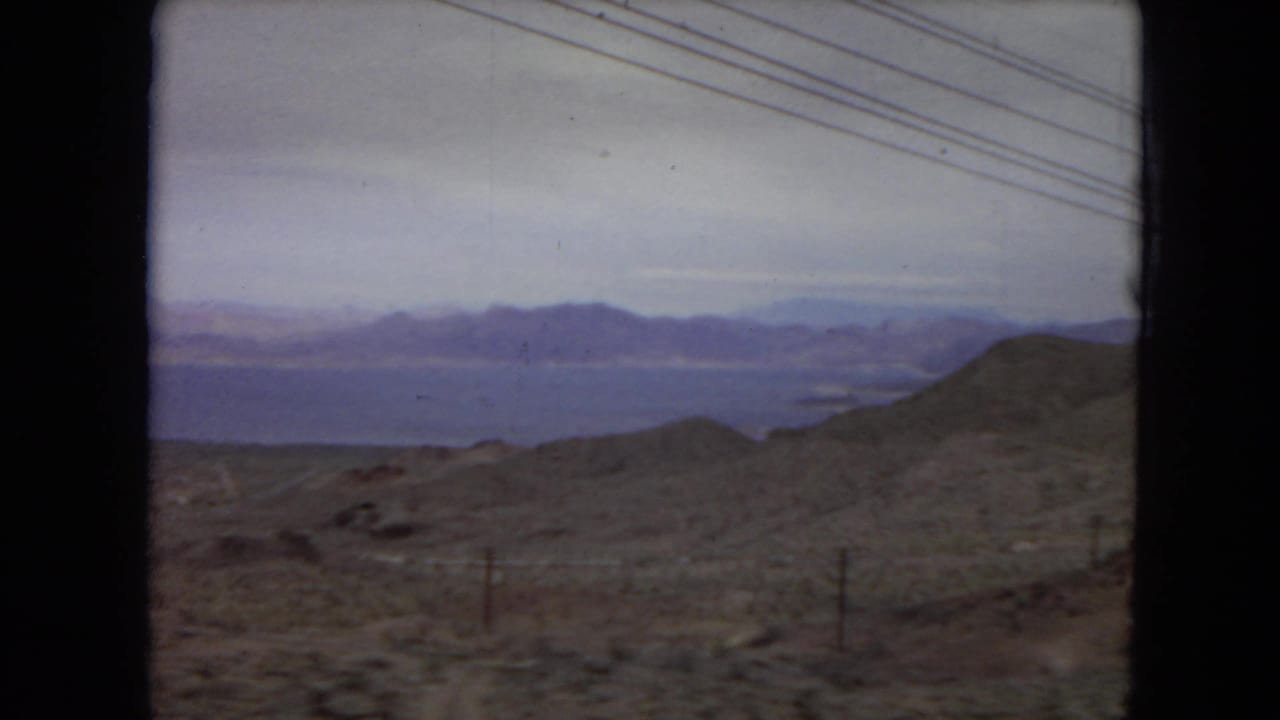 1967 United States travel frame from a moving viewpoint, with low desert hills, a distant blue water surface, mountain ranges, sparse fence posts, and utility lines.