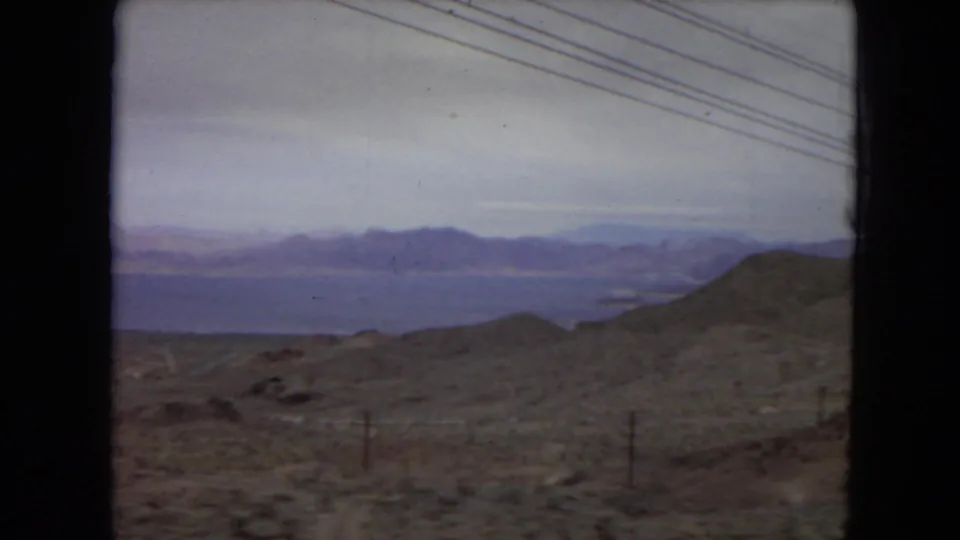 1967 United States travel frame from a moving viewpoint, with low desert hills, a distant blue water surface, mountain ranges, sparse fence posts, and utility lines.